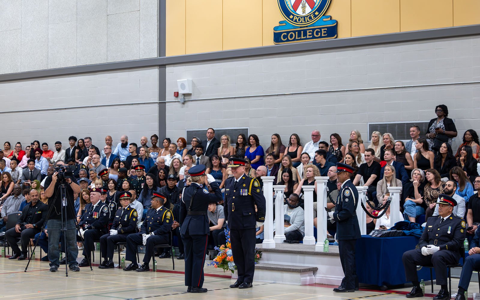 large group of people are seated inside a gymnasium hall, while a man and woman are standing, latter saluting to the former