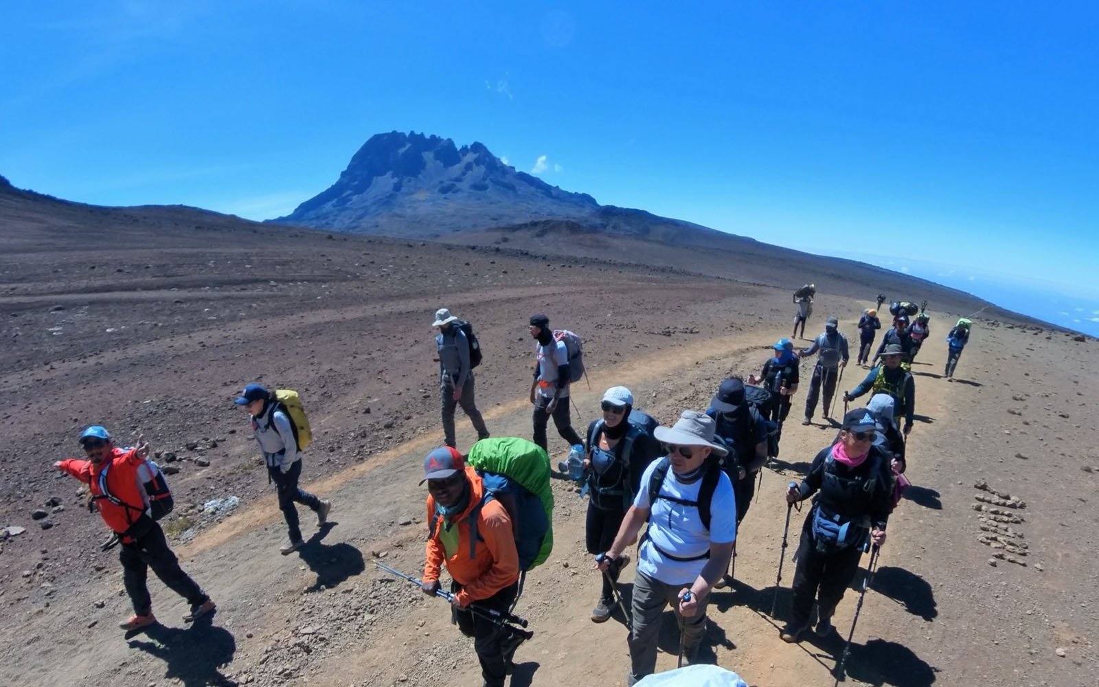 group of men and women walking along a dirt road with a tall mountain visible in the distance