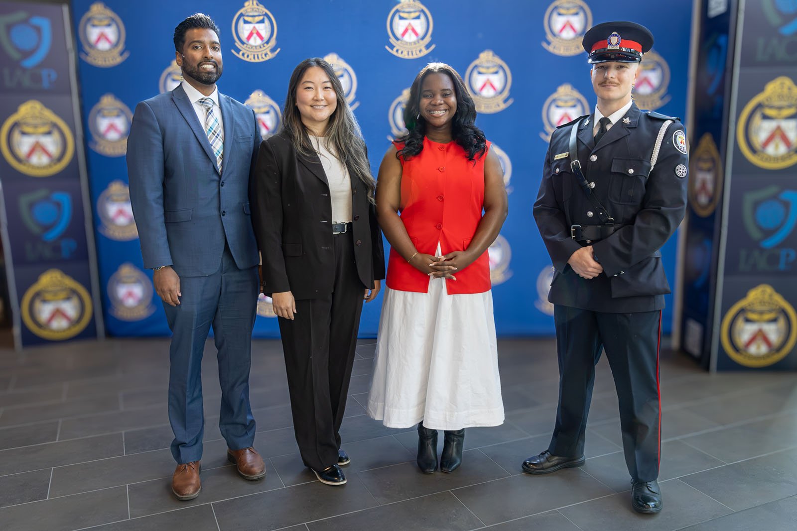 two women are flanked by two men posing for a photo in front of a backdrop with TPS crests on it