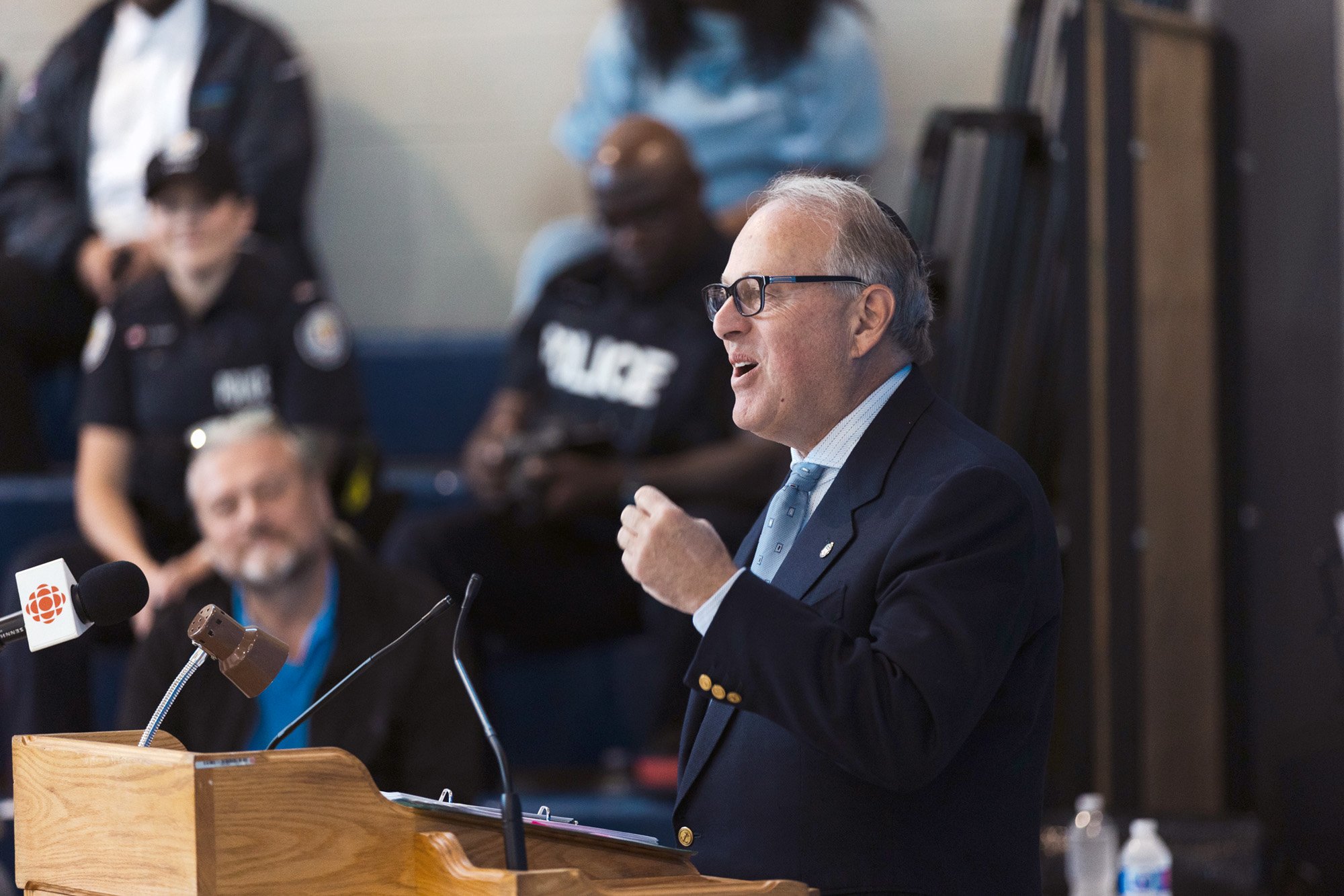 man with glasses dressed in a dark blue suit standing in front of a podium speaking
