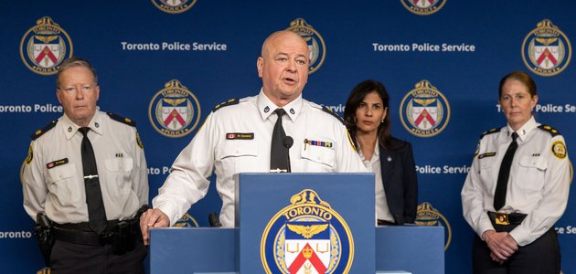man in a police uniform stands at a podium speaking while several other people are behind him listening