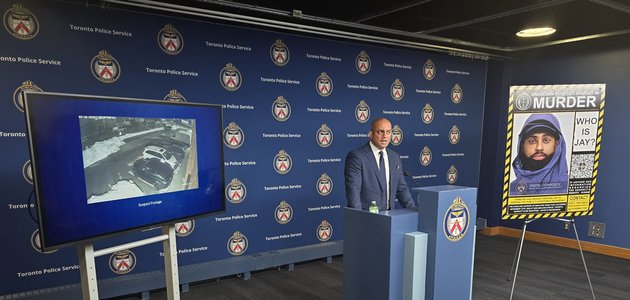 wide angle photo of a media room with a man in business suit standing in-front of a podium flanked by a TV screen on one side and a poster board on the other