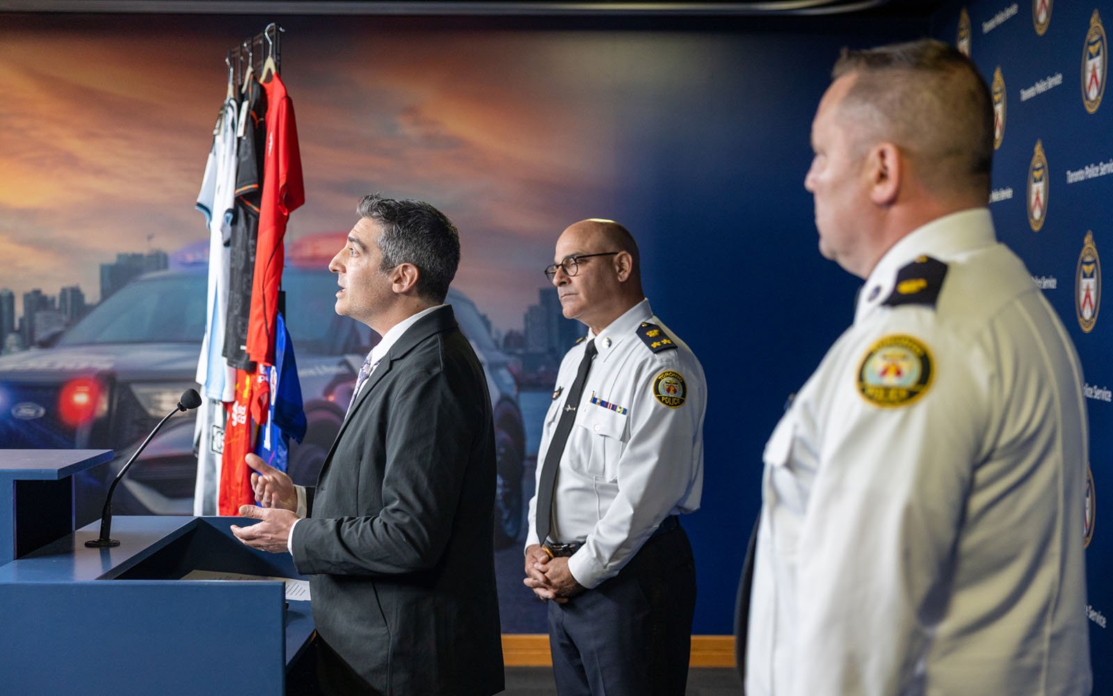 Side view of a man in a business jacket stands in front of a podium while two men in police uniforms stand behind him