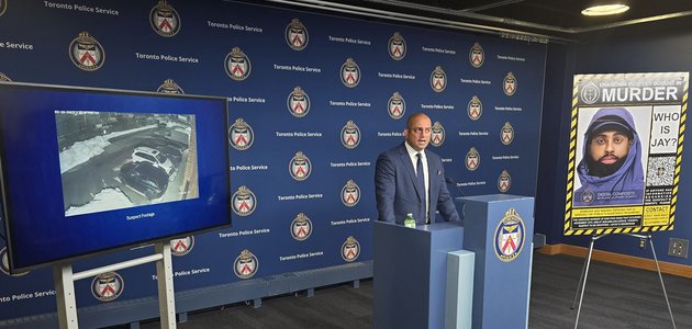 wide angle photo of a media room with a man in business suit standing in-front of a podium flanked by a TV screen on one side and a poster board on the other
