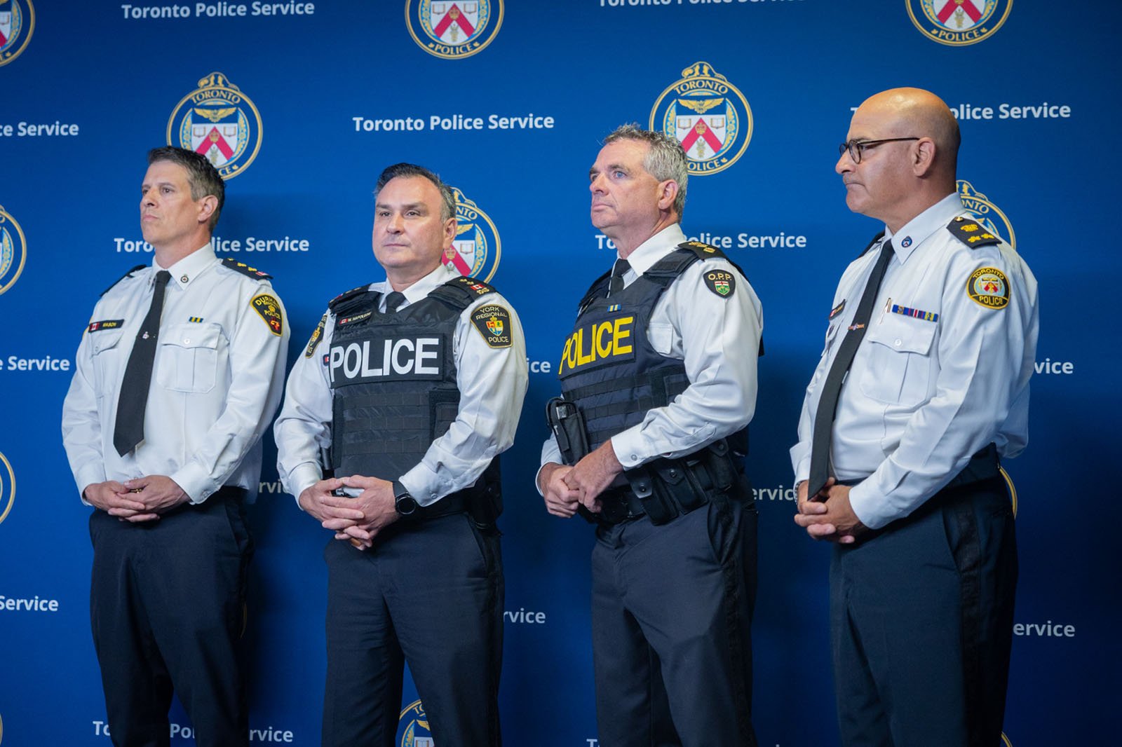 Four men in police uniforms stand in front of a wall that has a toronto police crest as a backdrop