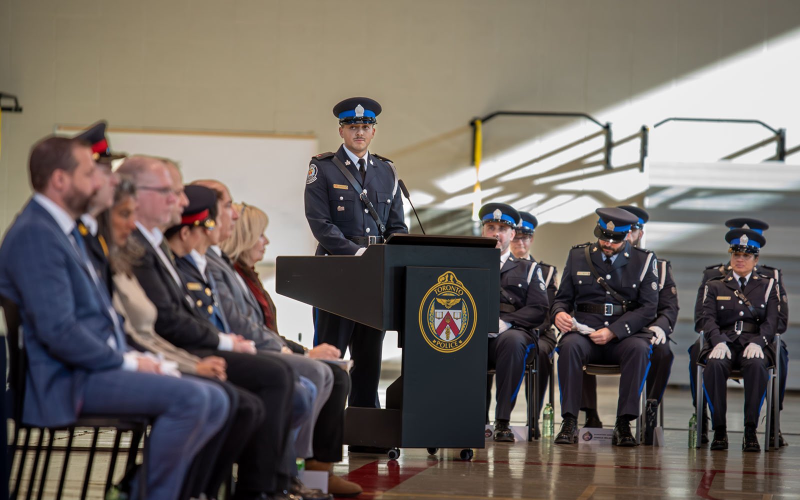 Man in a police uniform standing at a podium in a large hall with many other people sitting in the audience
