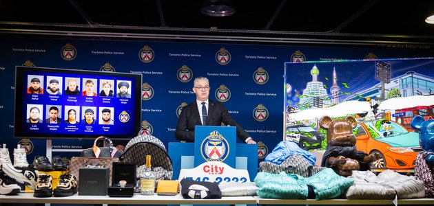 man in a suit standing at a podium next to a TV which is displaying a collage of photos, is surrouned by various electronics, luxury clothing, and other goods