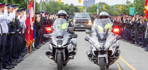 Two police motorcycles escort vehicles through two lines of saluting officers