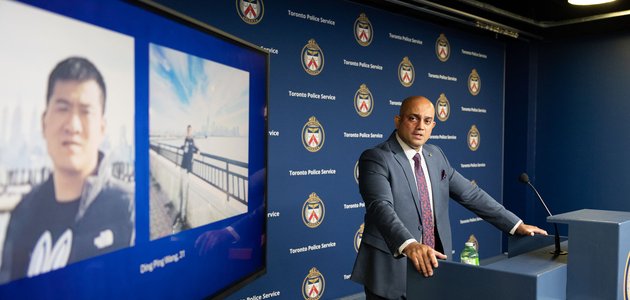 man at a podium looking at TV screen with aa photo of another man